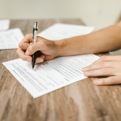 person filling out a paper form with a pen on a wooden table